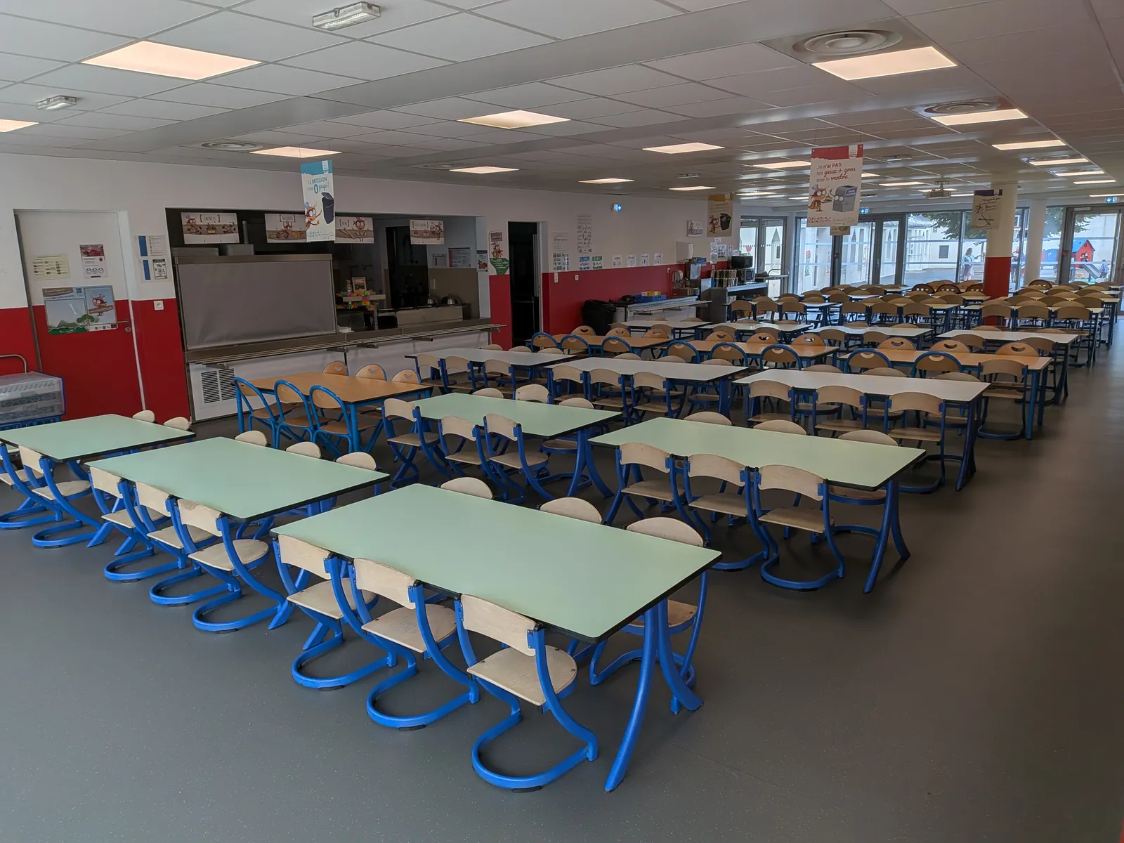 Salle vide aux longues tables vertes et chaises bleues alignées jusqu'aux baies vitrées