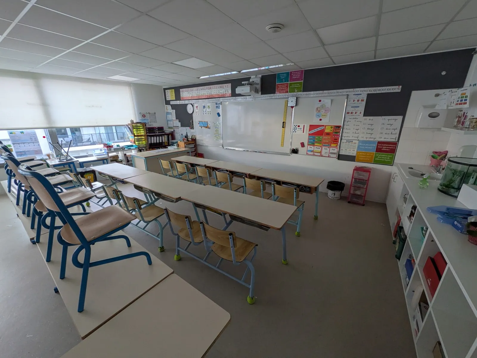 Salle vide avec des rangées de tables face au tableau blanc, des chaises posées sur les tables et des étagères de rangement le long du mur