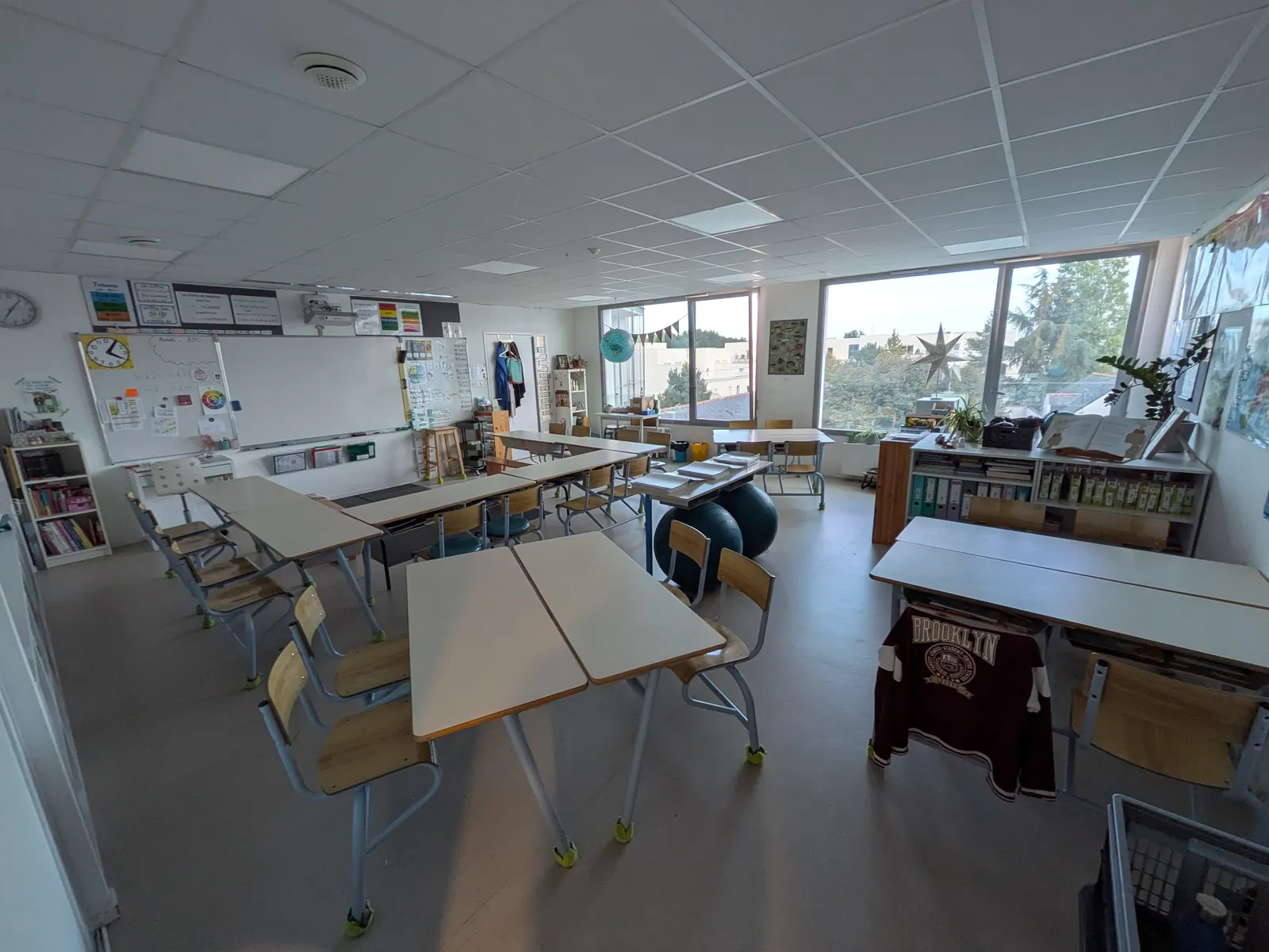 Salle éclairée par de grandes baies vitrées, avec des tables regroupées en îlots et des étagères de livres sous les fenêtres