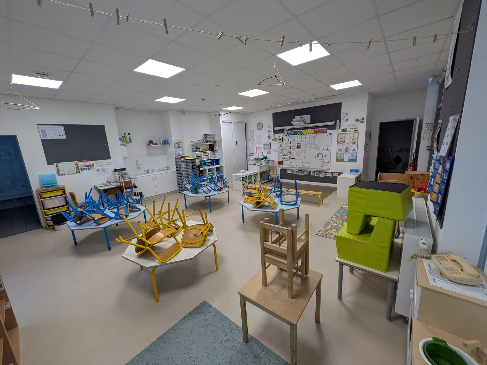 Salle vide aux tables basses, chaises retournées dessus, avec de gros blocs de motricité en mousse verte et des affichages colorés au mur